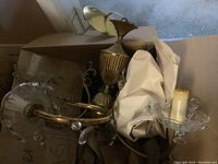 Two brass chandeliers placed in a box with packing paper, showing curved brass arms, some crystals, and candle sleeves on sockets. Crystals are missing in some areas.