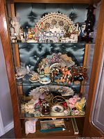 Full view of four shelves filled with a variety of porcelain and decorative figurines, hand fans and small collectibles inside a wooden and glass display case.