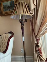 Full view of the floor lamp next to a cream upholstered chair and beige drapery with red tieback.
