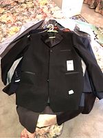 Photo showing multiple men's suit jackets stacked on a bed, the topmost being a black tuxedo jacket with satin lapels on a green hanger.