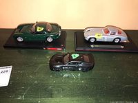 Three model cars shown together on a green surface, including a green TVR, a black muscle car, and a silver car with a yellow tape marked '10"'