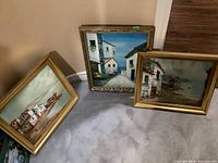 Three framed landscape paintings depicting coastal village scenes with houses and boats.