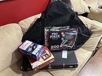 Poker accessories including two poker sets in cases, card shuffler box, and black carrying bag on beige sofa.
