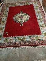 Full view of the fringed wool rug with red central field and detailed borders, laid out on a basement floor.