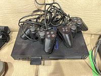 Front view of PlayStation 2 console with two wired controllers placed on top and memory card in console slot, showing PlayStation 2 logo and controller ports.