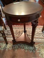 Front view of a round wood occasional table with a smooth top, drawer on the apron, three legs with carved angel heads, and a bottom shelf connecting the legs.