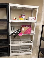 Photo of white plastic utility shelving unit showing five slatted shelves with various small items on them, all shelves and legs visible.