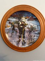 Circular framed painting of a winter scene with a boy skating on a frozen pond and other children in the background with snow and rural houses.