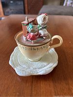 Front angle showing two mice dressed in winter clothing seated on the rim of a cream-colored teacup with green accents, with a fireplace and wrapped gift inside the cup, all mounted on a textured snow-like white plate base.
