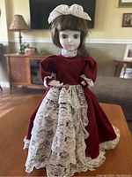 Front view of vintage musical doll featuring burgundy velvet dress with puffed sleeves, lace underskirt, lace collar, and white lace head bow; standing on wooden surface.