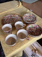 Photo showing complete set of 6 plates, 6 saucers, 4 teacups, milk jug, sugar bowl, and a larger plate with dense multicolor floral pattern on yellow fabric background.