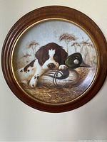 Plate with brown and white puppy lying next to two ducks and reeds in a round wooden frame.