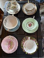 Top view of the six assorted vintage teacups and saucers arranged in two rows on a black tray, showing varied floral patterns and colors.