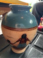 Front view of ceramic vase showing figure of person with hat and traditional clothing painted in earthy tones on beige background, upper portion glazed blue.
