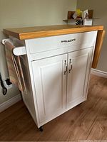 White wheeled microwave stand showing drawer on top, cupboard below with silver handles, and side tea towel holder with a hanging towel. Castor wheels visible underneath.