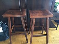 Two wooden bar stools side by side showing flat seats, leg design, and visible wear.