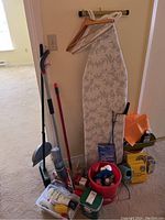 Photo of ironing board, mop, broom, dustpan, toilet auger, cleaning products, and plant pots arranged near a wall.