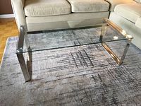 Full view of the glass and steel coffee table placed on a patterned carpet between cream-colored couches.