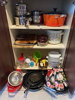 Cabinet shelf with stainless steel pots, orange enameled roasting pan, CorningWare casseroles, cutting boards, small crock pot, measuring cups, stoneware bean pot, frying pans and kitchen utensils.