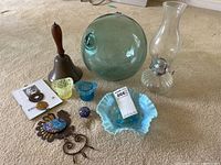 Full lot overview with all items including oil lamp, green glass orb, bell, small glass containers, blue ruffled dish, metal decorative pieces, and paperweight visible.