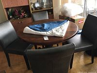 Round wooden dining table with four black leather-like chairs surrounding it. Blue tablecloth and white quilted table protector folded on table.