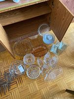 Overview of all glass servingware spread on floor in front of cabinet showing variety of plates, bowls, and platters