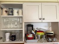 Photo of glasses and kitchenware including clear stemmed wine glasses, drinking glasses, plastic mixing bowls, measuring cups, and kitchen scale shown inside kitchen cabinets.