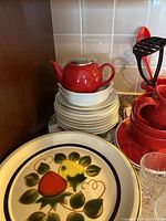 Photo showing stacked white plates topped with a small red ceramic teapot with metal strainer lid, next to larger red ceramic teapots and red kitchen utensils.