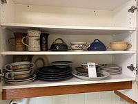 Photo of shelf with pottery mugs, covered dishes, bowls, and plates stacked.
