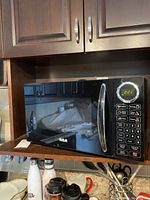 Black RCA compact microwave oven sitting on kitchen counter with cabinets above and various other kitchen items below.