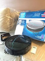 Robotic vacuum, charging dock, and original box seen from a distance on a beige surface.