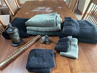 Towels of black and green shades neatly stacked alongside black bath mat, metal candle holder with cutout design, metal flower ornament, and gold-tone tie holder on wooden table.
