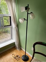 Black metal floor lamp with three white plastic shades positioned near a window with green wall background.