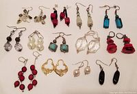 Overview of 13 pairs of assorted earrings in various styles, colors, and materials laid out on a white surface.