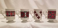Front view of the set showing four ceramic footed mugs with different Ukrainian embroidery style patterns in red, black, and white