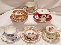 Six bone china teacups with saucers, various floral patterns and colors, arranged in loose grouping on white background.
