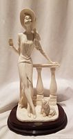 Full front view of vintage alabaster sculpture showing woman in hat with drink, leaning on balustrade with seated dog at base on wooden base.