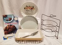 Full lot showing all items arranged: ceramic pie plate with apple pie design, fluted tart pan, rectangular baking dish, cookbook, metal racks, pie lifter, measuring spoons, and 3-tier pie plate holder