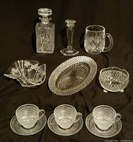 Full assortment of crystal and cut glass items including decanter, candlestick, mug, three serving bowls, three teacups and saucers on black background.