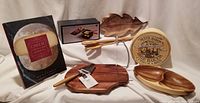 Photo showing assorted wood cheese boards and trays, Brie Baker box, cheese book, and Raclette melter items arranged on fabric.