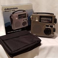 Photo showing the Grundig FR200 emergency radio, its original box, and black carrying case displayed on white surface.