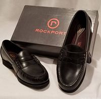 Pair of black men's Rockport leather shoes shown slanted with original box behind.