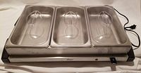Front view showing three stainless steel buffet pans with clear plastic lids placed in warming tray base, power cord visible.