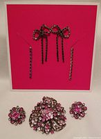 Photo showing large pink rhinestone floral brooch with two matching pink rhinestone buttons and two pairs of earrings (black bow-shaped and lilac-colored line) displayed on a bright pink background.