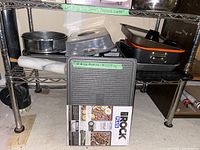 Wide view of assorted baking and roasting pans including springform pans, a rolling pin, The Rock Plus grill/griddle in box, and various other pans on shelves