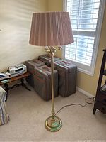 Full view of tall brass floor lamp with pleated shade standing in a corner of a room, near window and luggage.