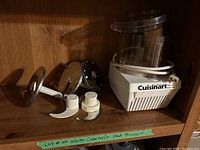 Photo of white Cuisinart food processor base with clear work bowl, feed chute, and various attachments including dough hook, slicing blades, and grating disc on wooden shelf.