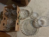 Full view of assorted cut glass bowls, plates, and bottles grouped on carpet and in cardboard box.