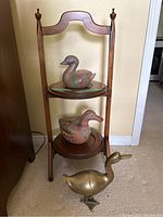 Full view of wooden stand holding two ceramic ducks and brass duck placed on floor beside it