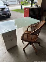Desk with heavy glass top on two white drawer column pedestals and wooden desk chair with casters positioned nearby.
